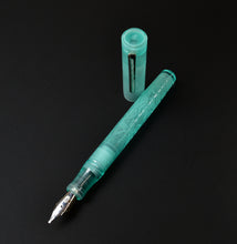 Load image into Gallery viewer, A translucent mint green acrylic named sea glass, fountain pen with floral engravings on the barrel, shown with its matching cap standing upright on a solid black background.