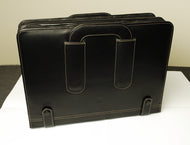 A black leather briefcase stands upright beside a Franklin-Christoph 80 Pen Case on a two-tone surface.