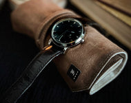 A Franklin-Christoph IWO Timepiece with a black leather strap sits on a brown suede roll next to a closed book.