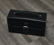 Black leather briefcase with handle and buckle, next to Franklin-Christoph The Christoph 48 Pen Case on wood floor.