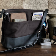 A Franklin-Christoph Fortis Sling Bag - Iron Brown and Black rests on a desk with maps, wallets, and a vintage vase.